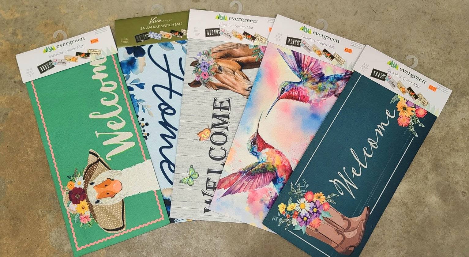 Collection of colorful 'Welcome' door mats with various designs on a beige surface.
